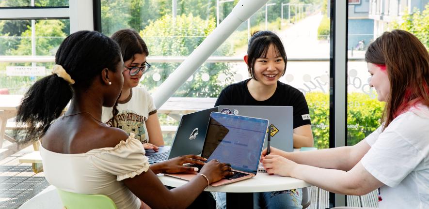 A group of University of Cambridge students working on laptops around a small table in front of a large window at the West Café. Copyright: University of Cambridge (June 2023)