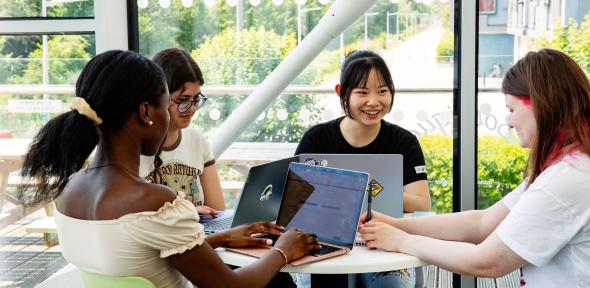 A group of University of Cambridge students working on laptops around a small table in front of a large window at the West Café. Copyright: University of Cambridge (June 2023)